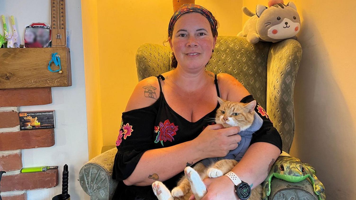 Morgan the cat wearing a grey jumper being held by his owner Rhia, who is sitting on a living room chair