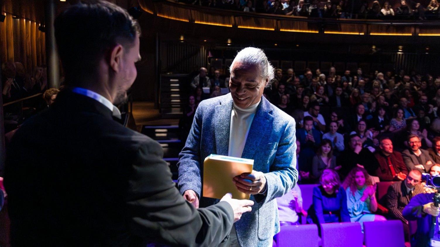 Mike Elliot accepting his award on stage. He is wearing a white turtleneck jumper underneath a grey tweed blazer, with grey trousers. He has long, dark grey hair tied back into a slick low bun. He is holding the award in one hand and shaking the hand of the presenter with his other. The presenter is facing away from the camera.