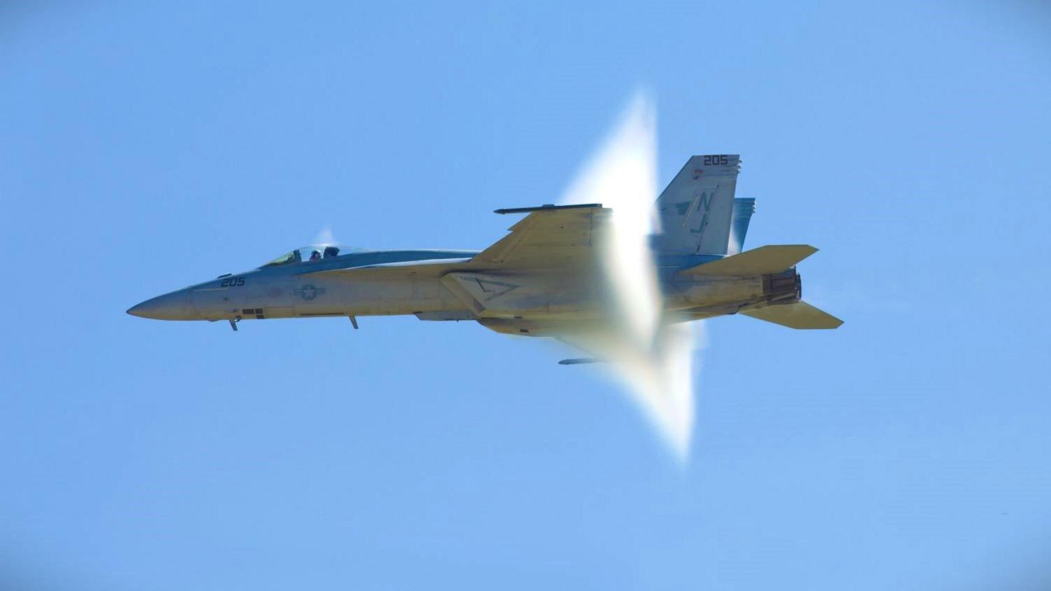 A grey fighter jet flying across a blue sky and creating a cloud-like pressure wave towards its tail.