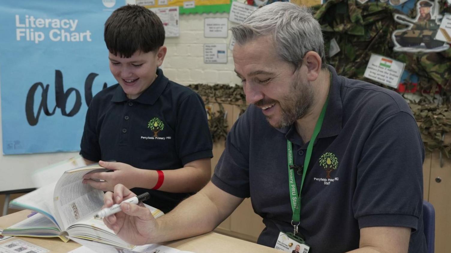 Jacob, wearing a navy polo shirt with the Perryfields logo on it, smiles as he turns the page of a workbook. He is sitting next to a member of staff who has grey hair and is holding a black marker pen.