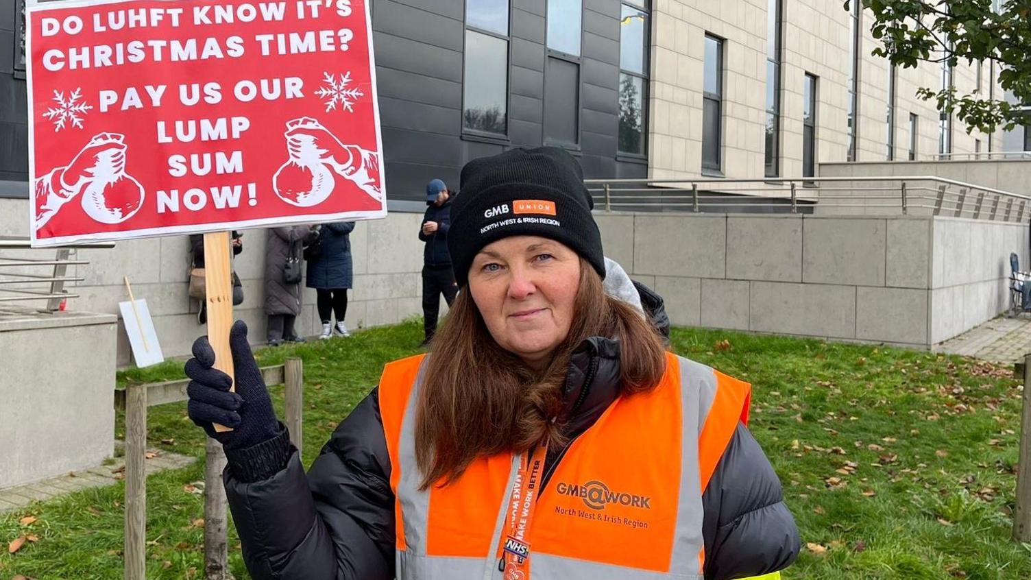 Kerry Nash, a senior organiser at the GMB union, stands on the picket line outside Royal Liverpool University Hospital holding a placard with the words 'Do LUHFT' know it's Christmas time? Pay us our lump sum now!'