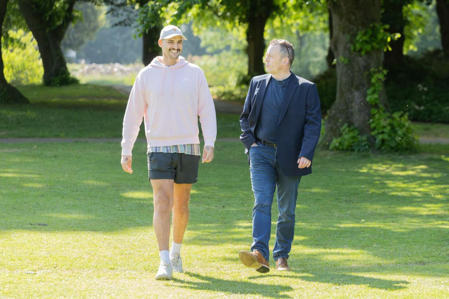 Two men walking on grass in a park on a sunny day. On the left is a tall man wearing a light-pink hoodie, a white baseball cap and shorts. He has a dark moustache and is smiling broadly. On the right, an older man is wearing a blue blazer and blue jeans and is looking at the other man.