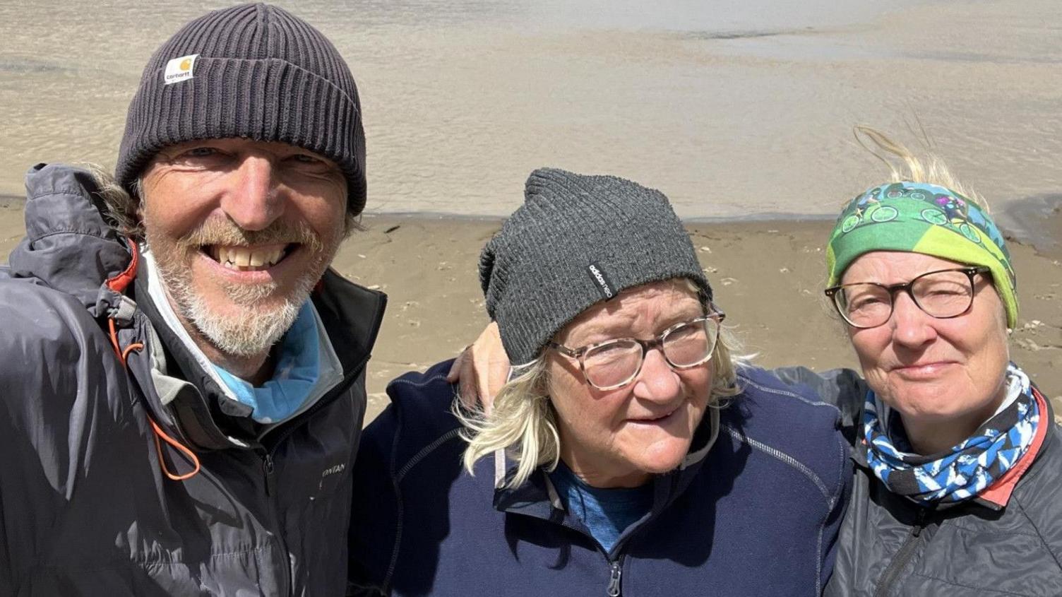 Craig, Sophie and Rona are in the outdoors wearing and smiling for a picture Craig is taking on his phone. Craig and Sophie are wearing fleece hillwalking hats while Rona has colourful scarf around her head.