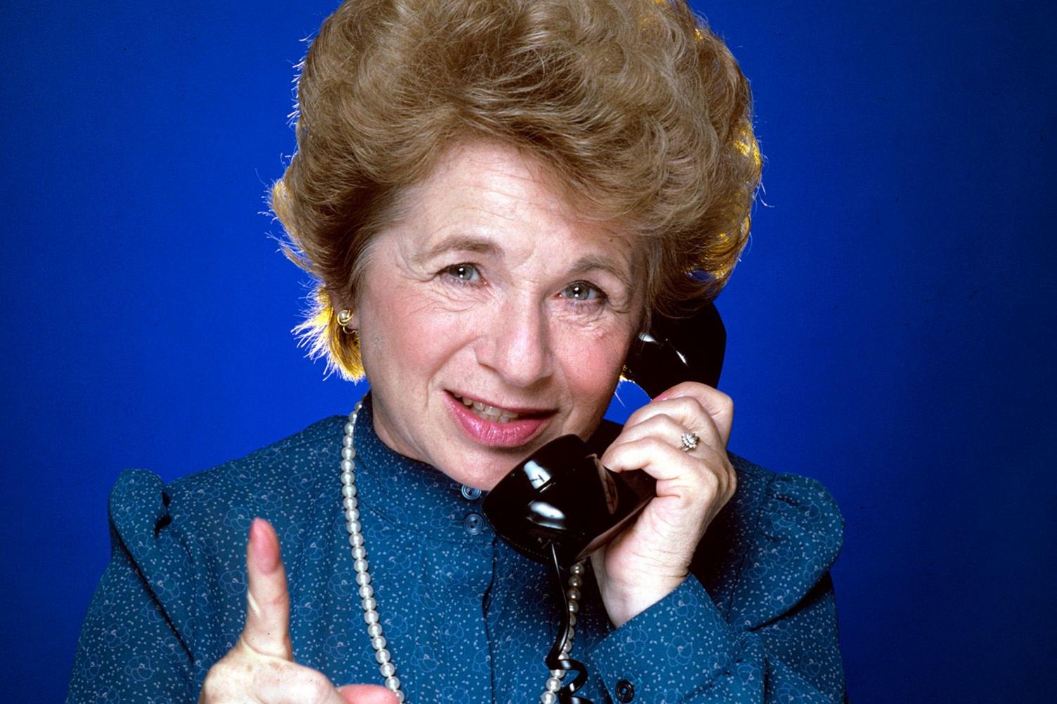 Publicity shot of Dr Ruth Westheimer, pretending to be on the phone and presumably giving advice to a phone-in caller. She is looking straight at the camera meanwhile and waving a finger. She has short styled chestnut hair and is wearing a blue collarless blouse buttoned to the top, and a string of pearls