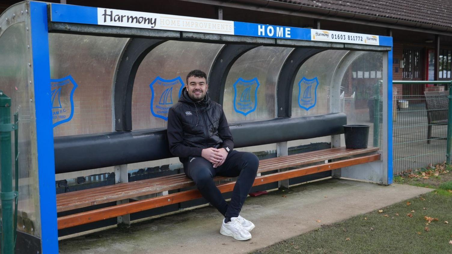 Wroxham midfielder Jordan King wearing black sportswear and smiling. He's sitting in the club's dugout.