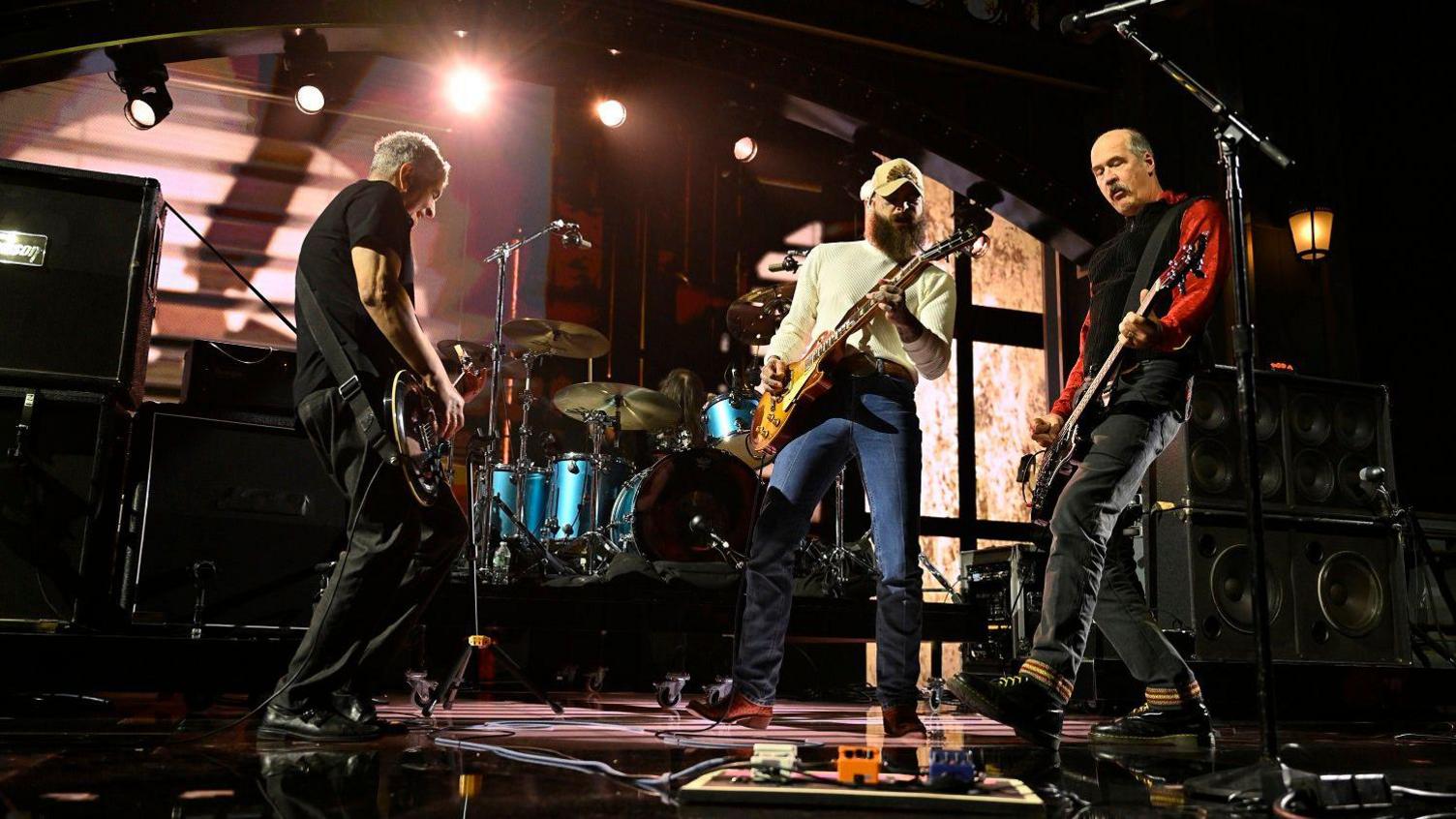 Three men perform on stage with guitars with a drum kit behind them