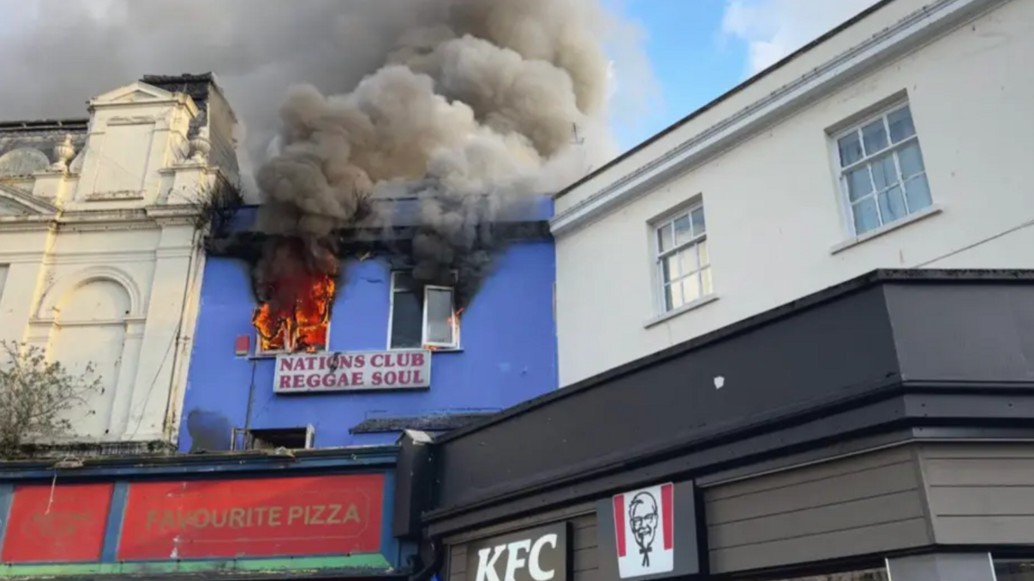 Flames and smoke pour out of the upstairs windows of Nations Club on Union Street. The building is blue and is above a pizza shop. In the bottom of the image a KFC logo can be seen, which is the building next door.