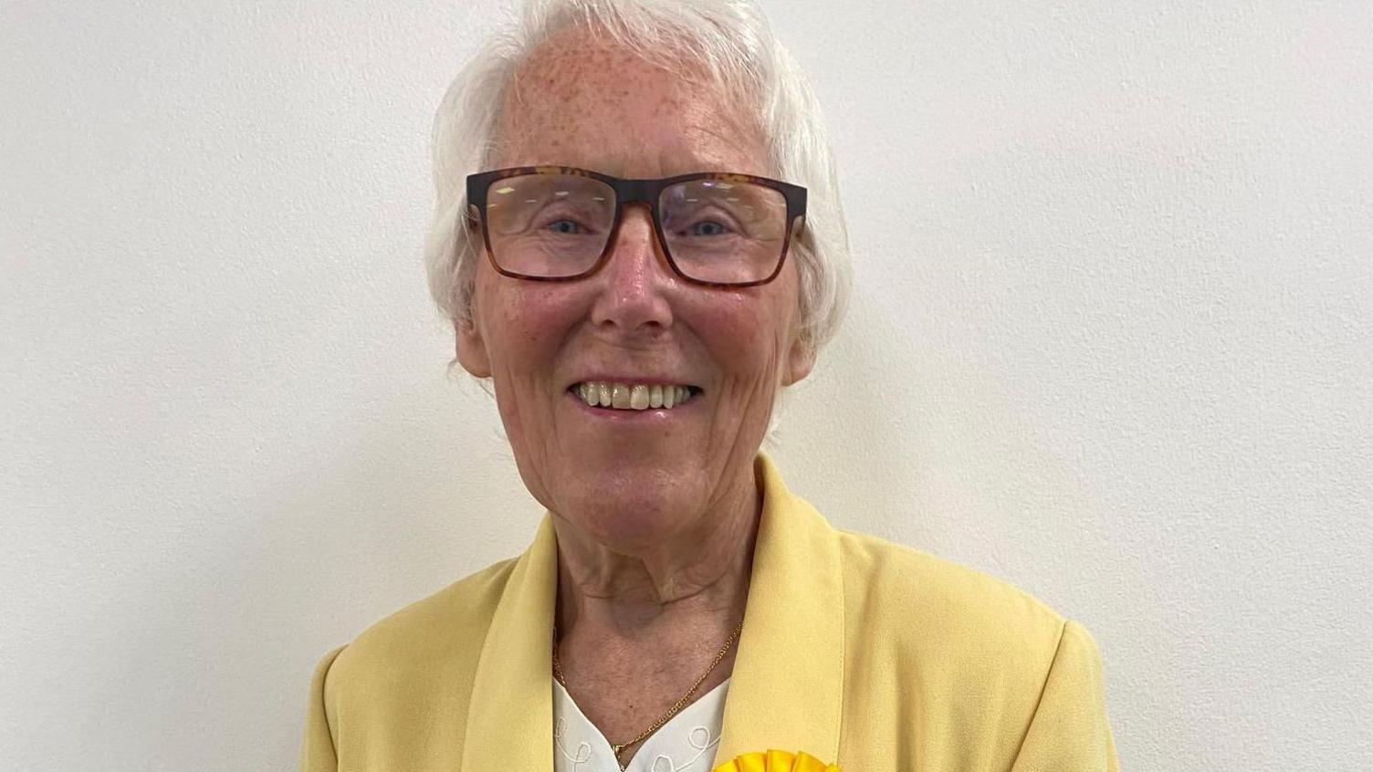 A smiling woman wearing glasses with short, grey hair. She is wearing a cream top underneath a yellow jacket with the top of a yellow rosette showing. 