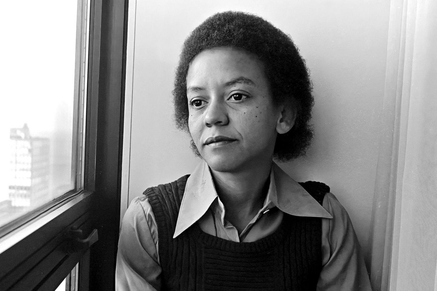 A black-and-white photo of Nikki Giovanni in a hotel room in Chicago in the mid-70s. She is looking thoughtfully out of a window on an upper floor. She has shortish hair and is wearing a black tank top over a shirt with wing-tip collars