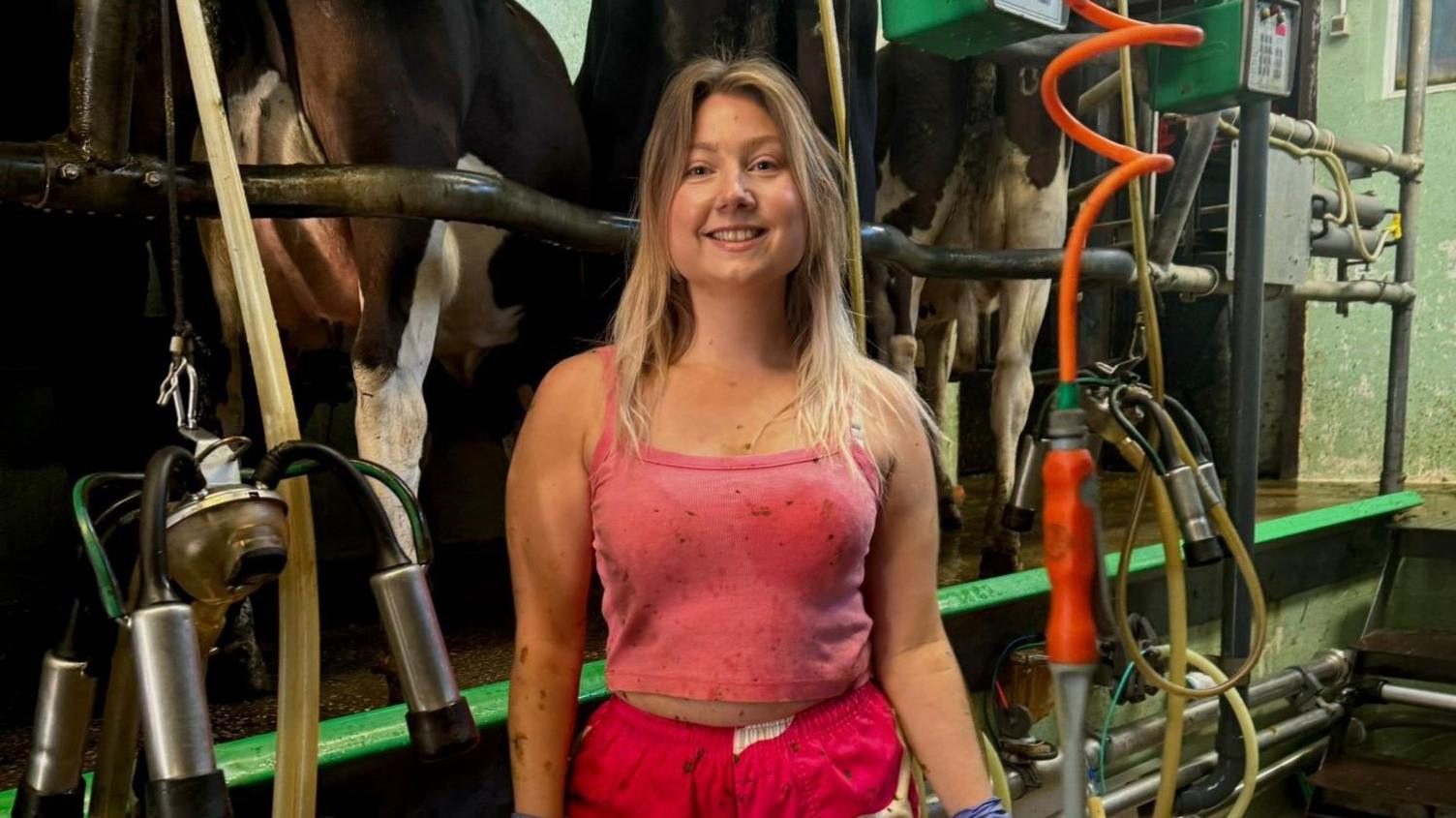 Lizzie McLaughlin is standing in front of a milking machine. She's a young woman wearing a pink top and red trousers. She has long blonde hair 