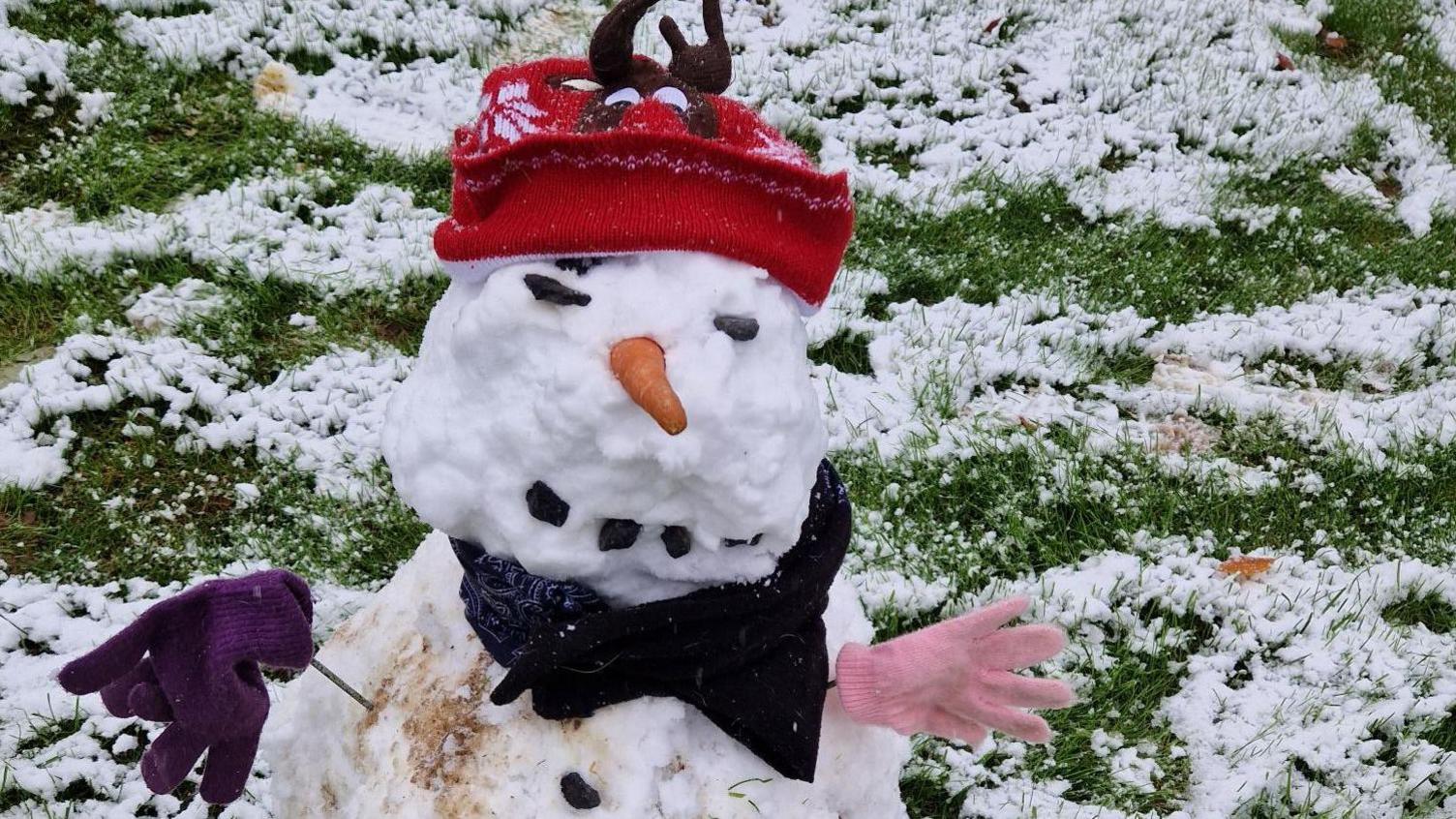 A snowman is sitting on some grass on Exmoor. He is wearing a red hat and two mismatched gloves. He has a carrot as a nose.