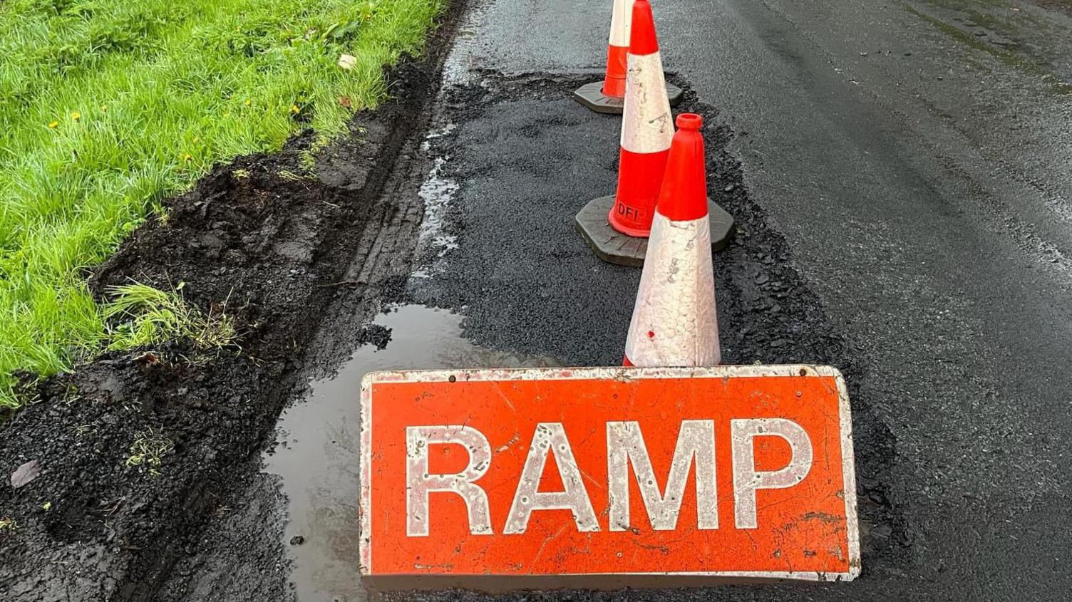 A road with a massive pothole, grass on the left with thick mud and a fluorescent orange sign that says 'RAMP' in capital letters on it in front of fluorescent orange and white cones. 