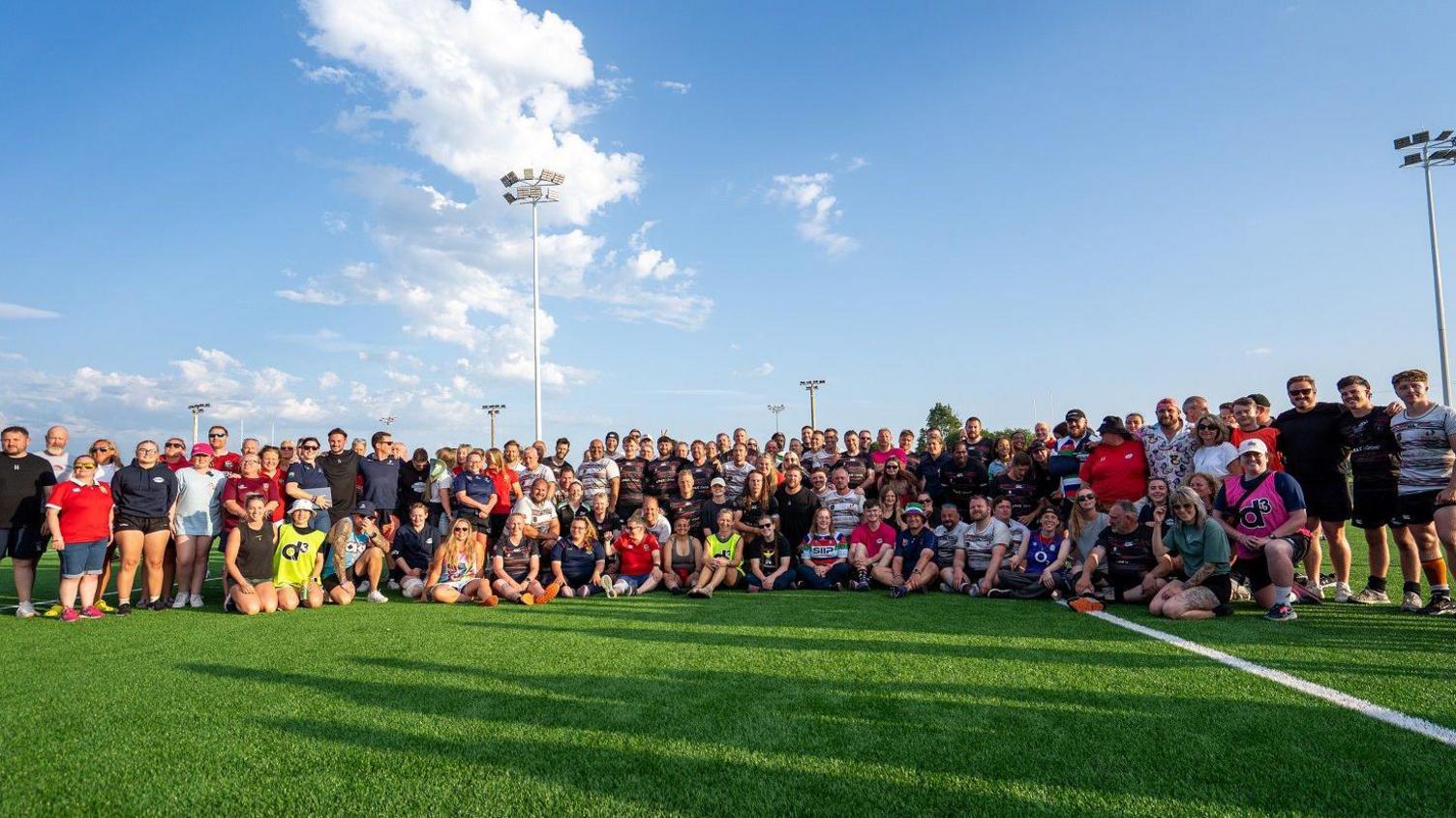Around 50 people wearing rugby kits posing for a photo on a sports pitch. It's a sunny day and the sky is blue.