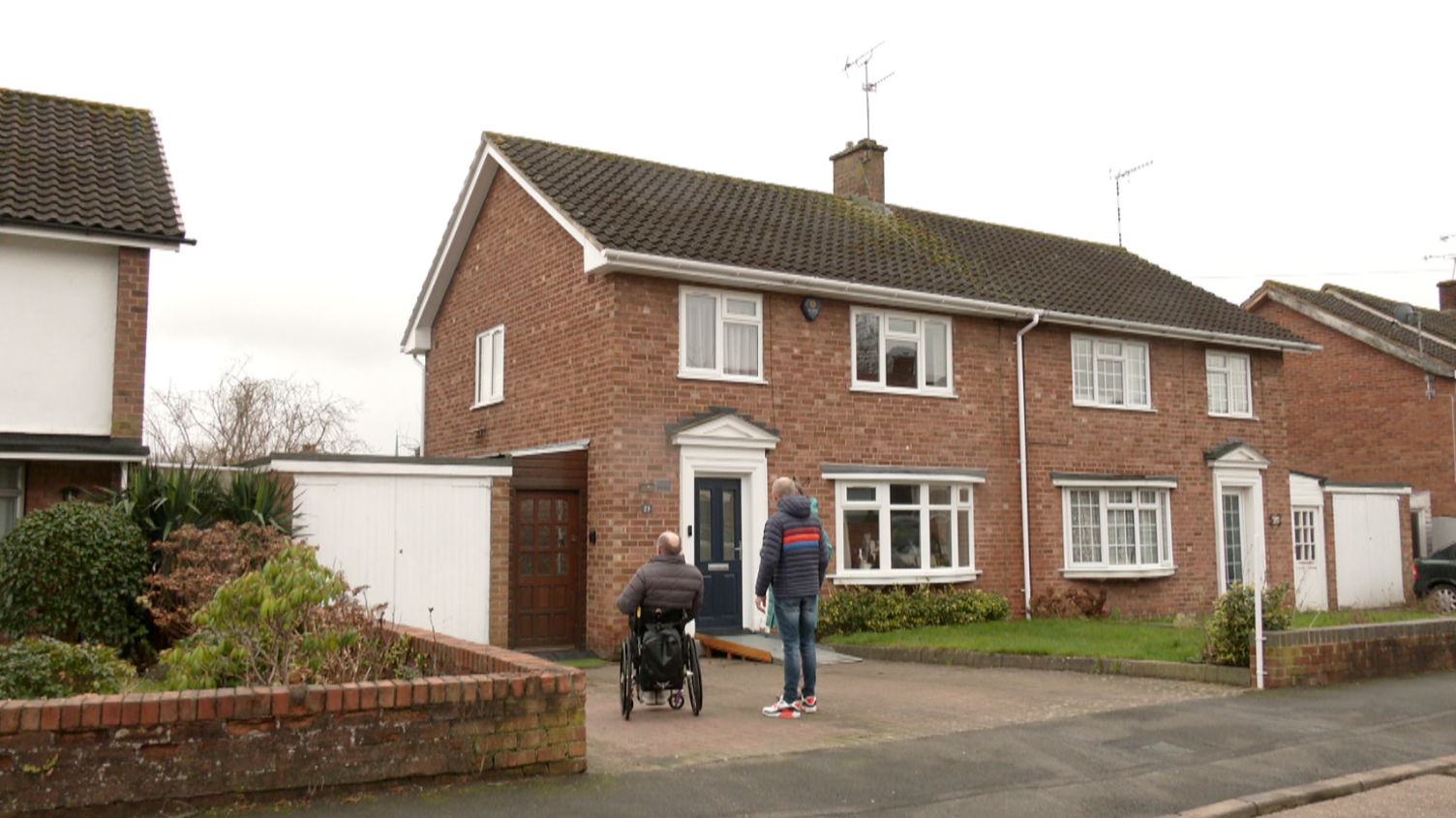 Two men, one in a wheelchair and one standing facing a semi-detached brick house. There are properties either side.