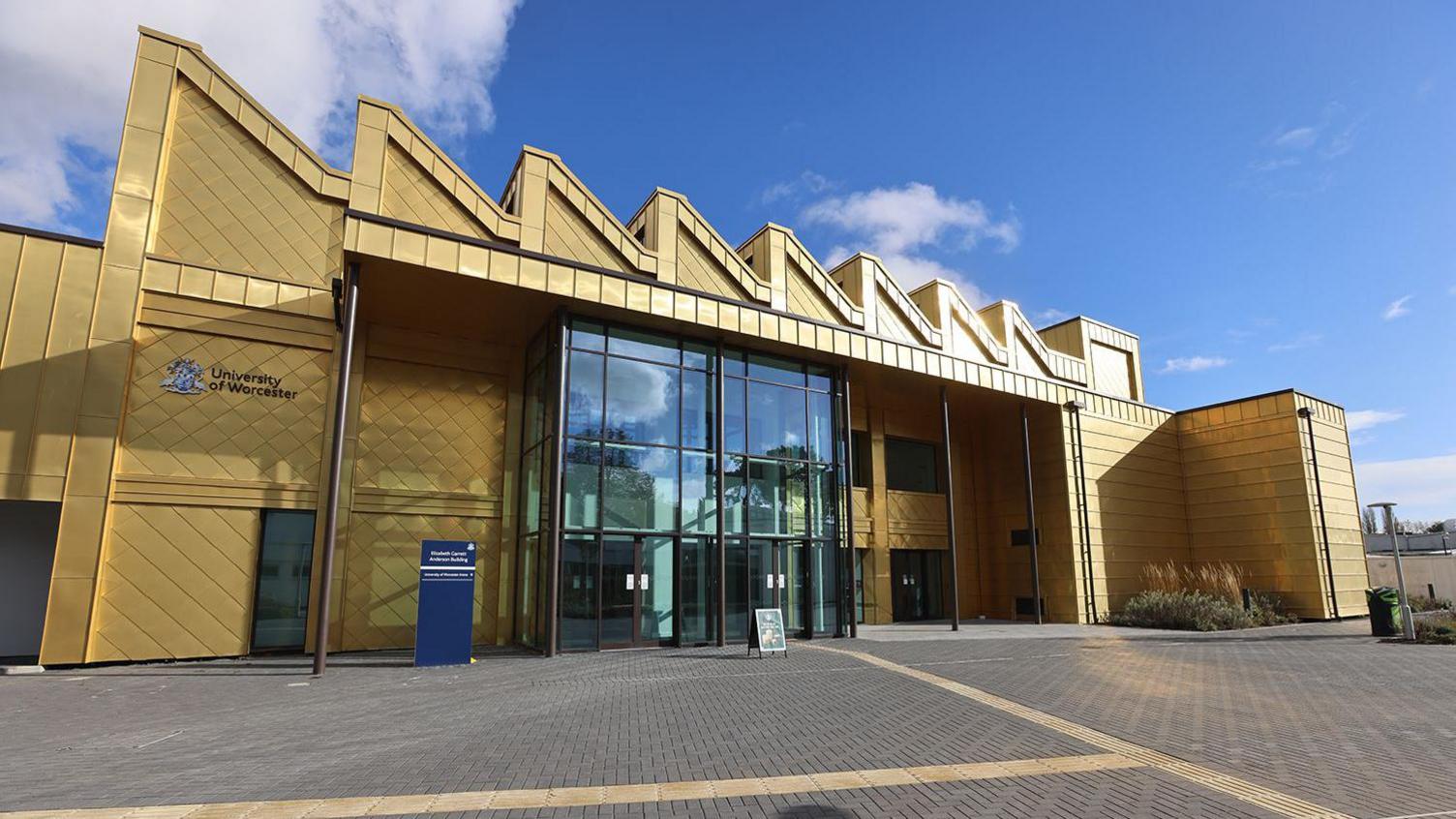 A multi-storey building with a shiny gold exterior and a large glass entrance is photographed on a sunny and largely clear day.
