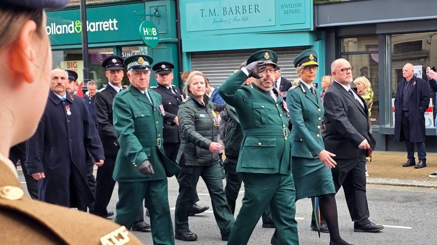 NHS workers in green uniforms march in formation down a high street, all looking to the right where a woman stands in a brown military uniform.