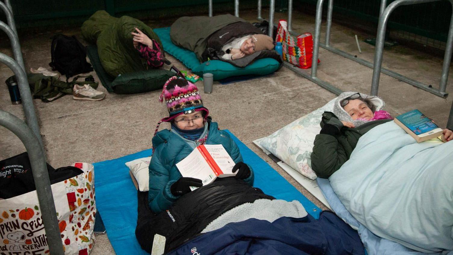People in sleeping bags wrapped up warm and smiling at the camera. It is a cold looking night and a couple of the women hold books.