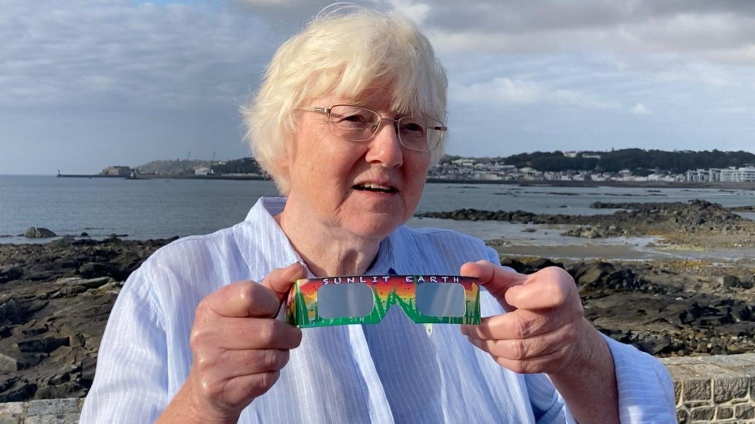 A picture of Dr Jean Dean. She is wearing a blue shirt and is holding a pair pf carboard glasses as she stands with the sea behind her.