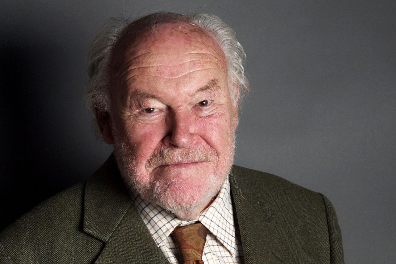 A photo of Timothy West taken when he was a fairly elderly man, standing against a grey background. He is mostly bald, with some grey hair and is unshaven. He is wearing a greyish jacket, a checked shirt and a floral tie.