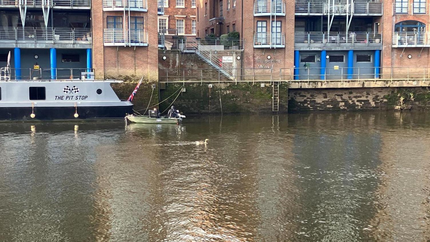 A stretch of river with a search boat on the water. There are buildings in the backdrop overlooking the river.