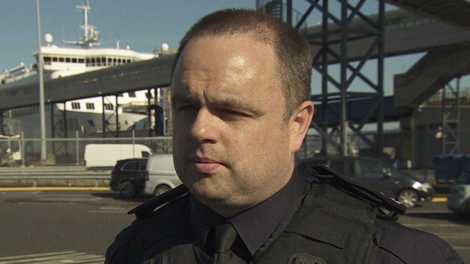 Johnny Evans - a man with short hair wearing a dark Immigration Enforcement officer uniform looks beyond the camera. He is standing in the middle of a car park with a large boat in the background.