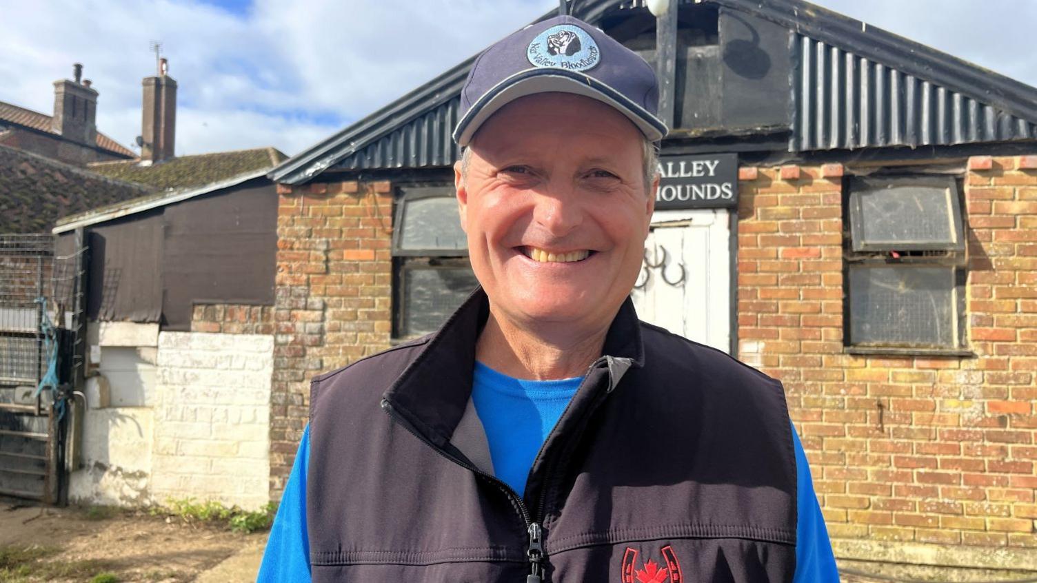 David Hunter wearing a black gilet and blue baseball cap and standing in front of a brick putbuilding