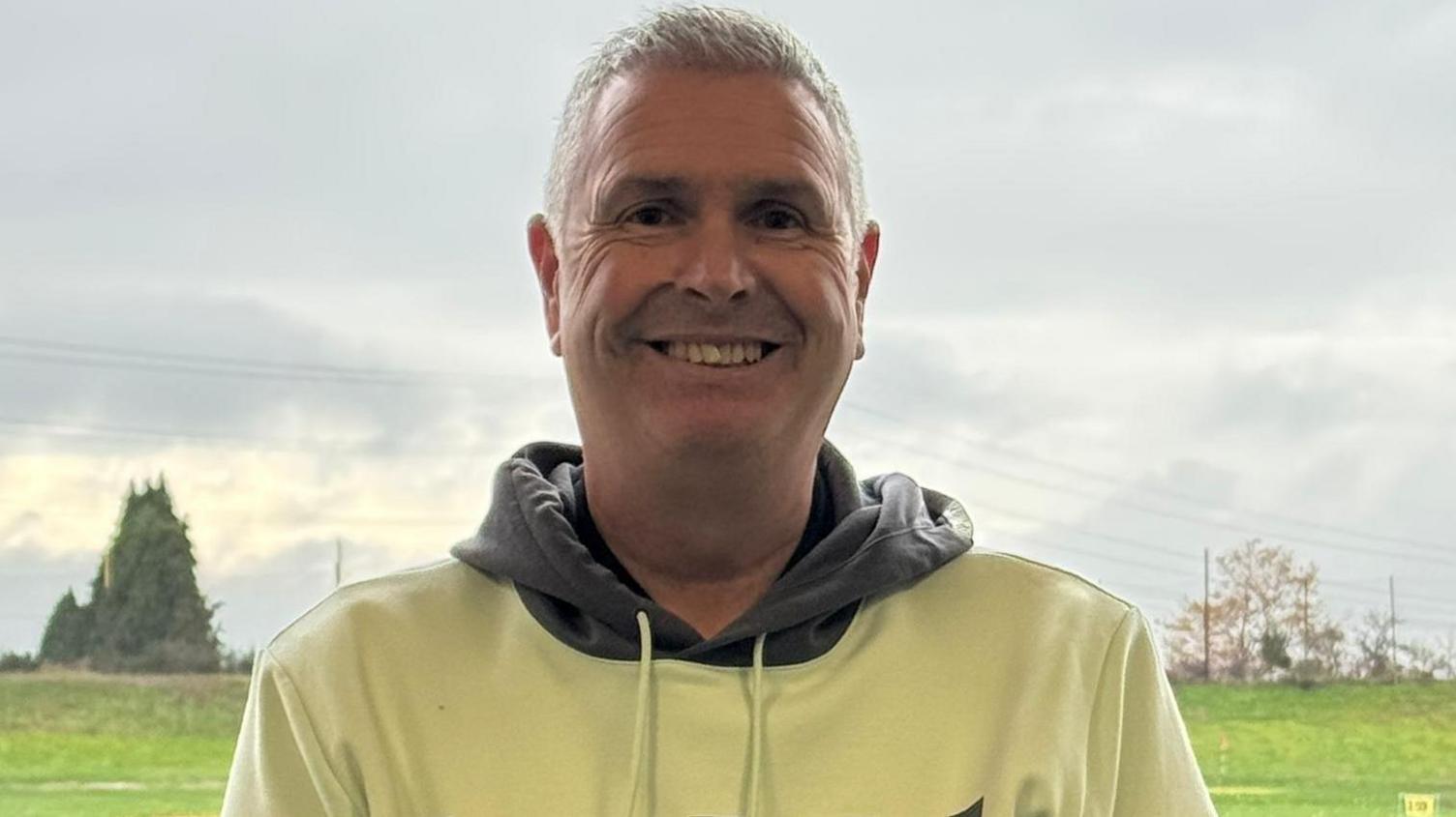 Darren is wearing a cream hooded top with a grey hood. He is smiling at the camera and has cropped grey hair.