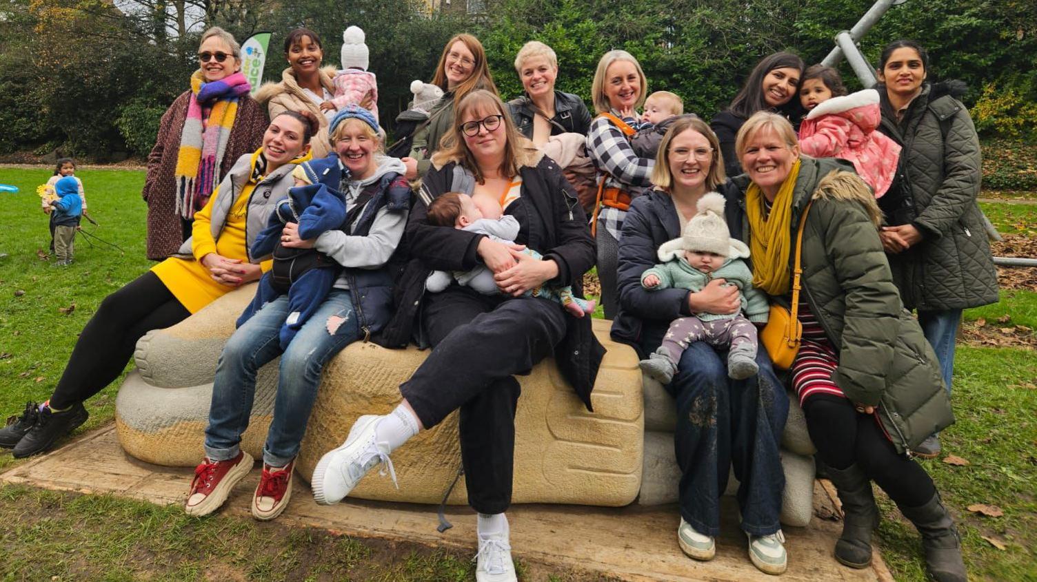 A group of women, some holding babies, sitting on a stone sculpture in a park