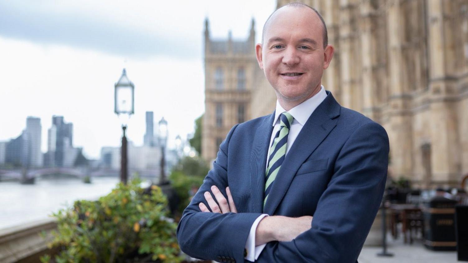 A picture of Fylde MP Andrew Snowden taken with the Palace of Westminster to the right of him and the River Thames to the left. He's wearing a blue suit and stripy pale green-and-navy tie. He has his arms folded in front of him and is smiling for the camera.
