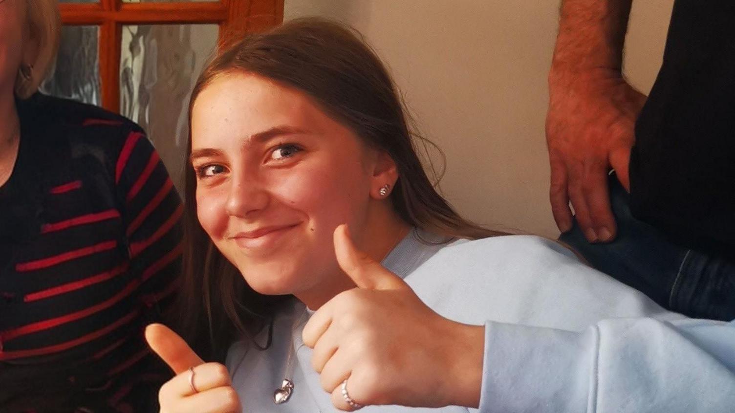 Chloe Longster puts both of her thumbs up in this picture looking directly at the camera. She is wearing stud earrings and is smiling, she has long, straight, brown hair and is wearing a silver necklace. She has a white sweatshirt on.