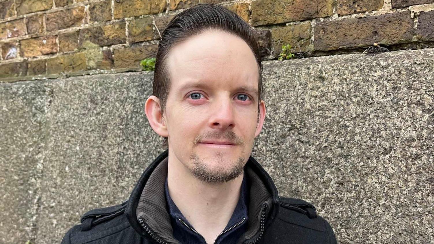 Dan Oakley stands next to Sheerness Dockyard wall. He has a black coat and is looking directly at the camera. 