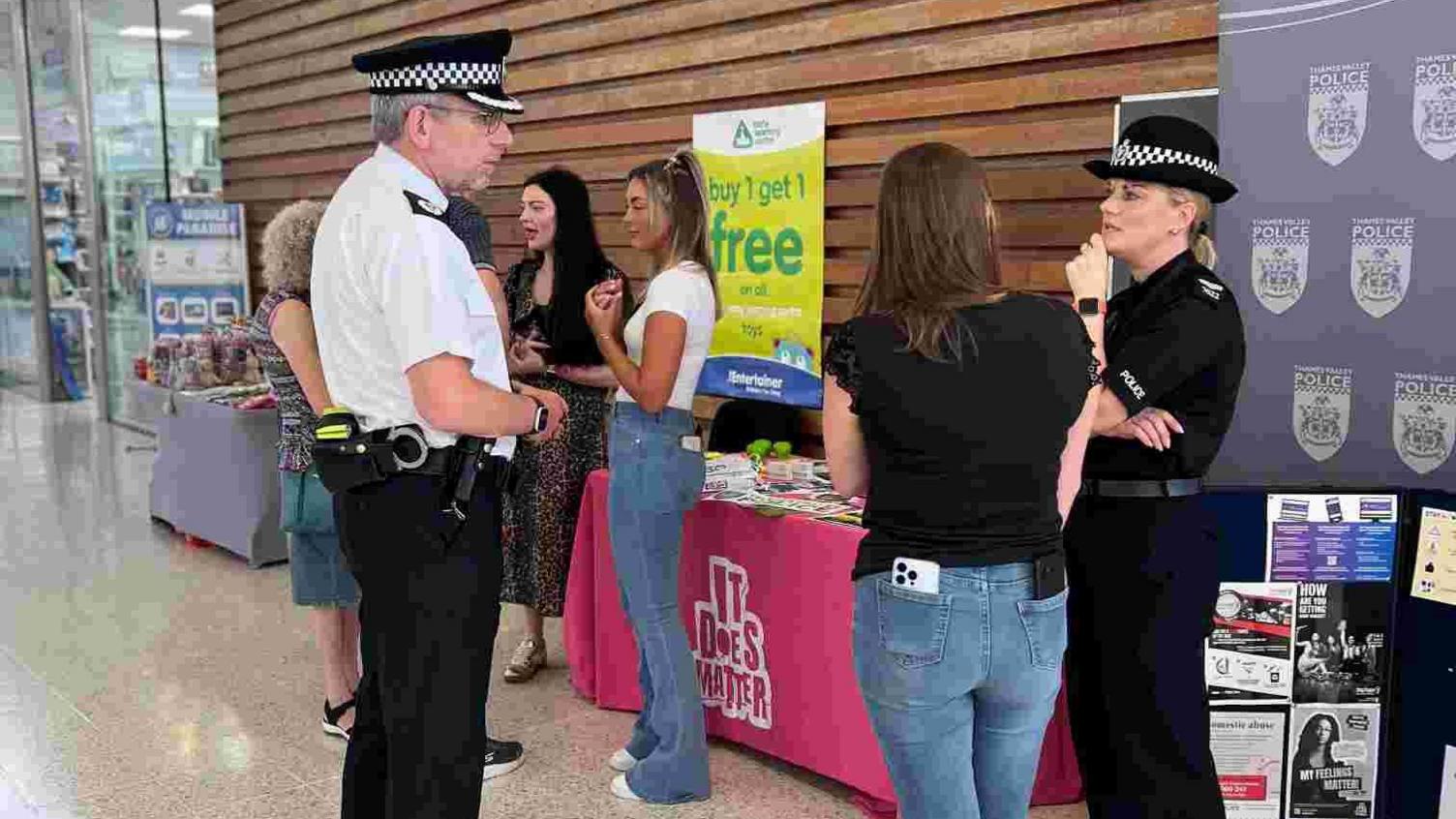 Two police officers, talking to a woman, with her back to the camera, and other women in the background. The male officer is looking at a female officer, and they are side on. They are both in uniform, with hats on. There are stalls behind them with leaflets and information about domestic violence. 