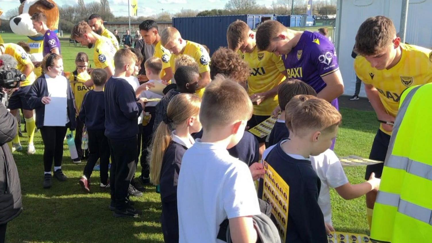 A wide shot of a row of Oxford United players, wearing a mix of yellow and purple kits, signing pieces of paper for a crowd of Year 6 students.