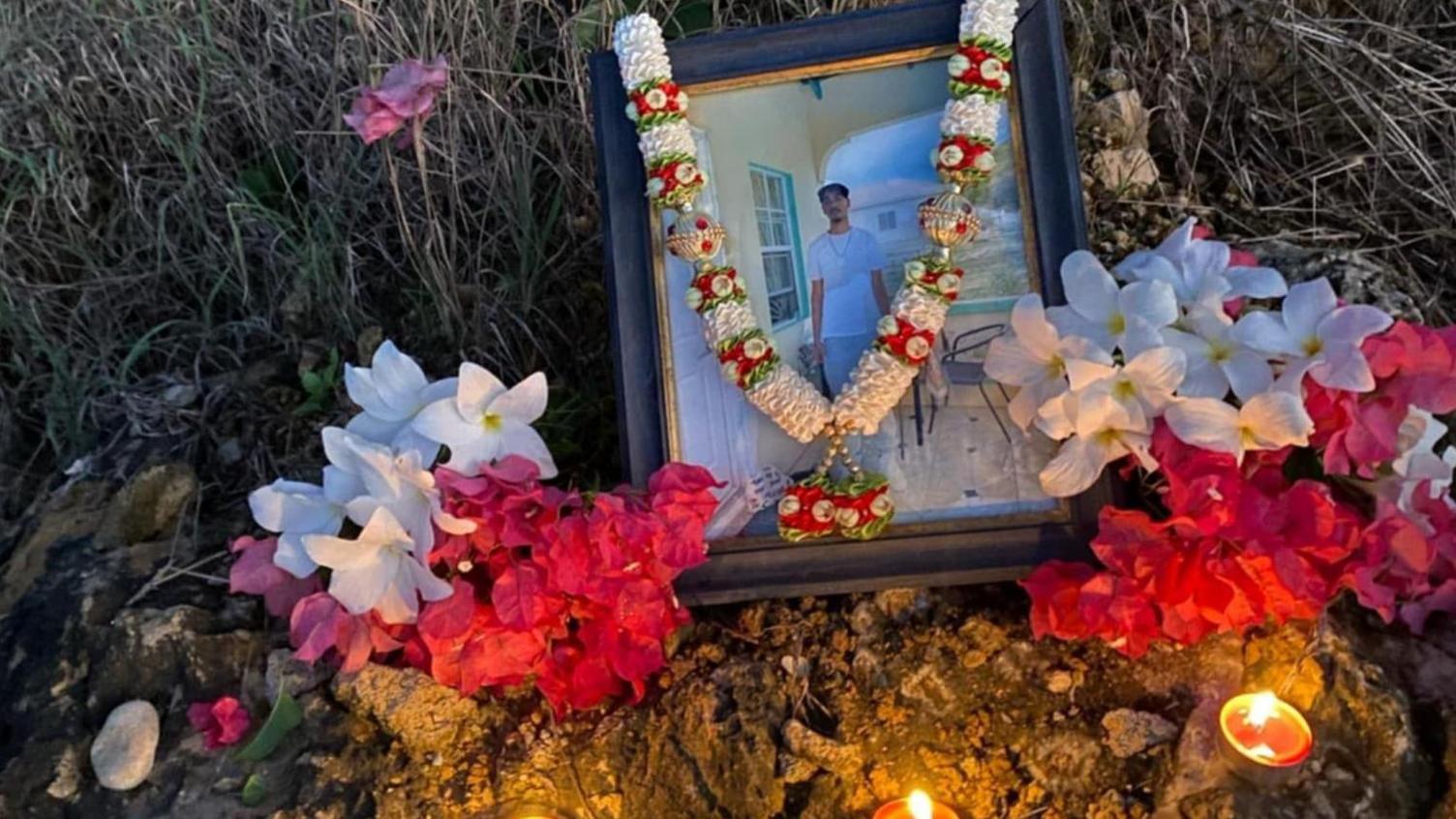 A photo in a frame of Shawn sits on the ground by the beach. It has a floral garland around it with flowers and candles next to it.