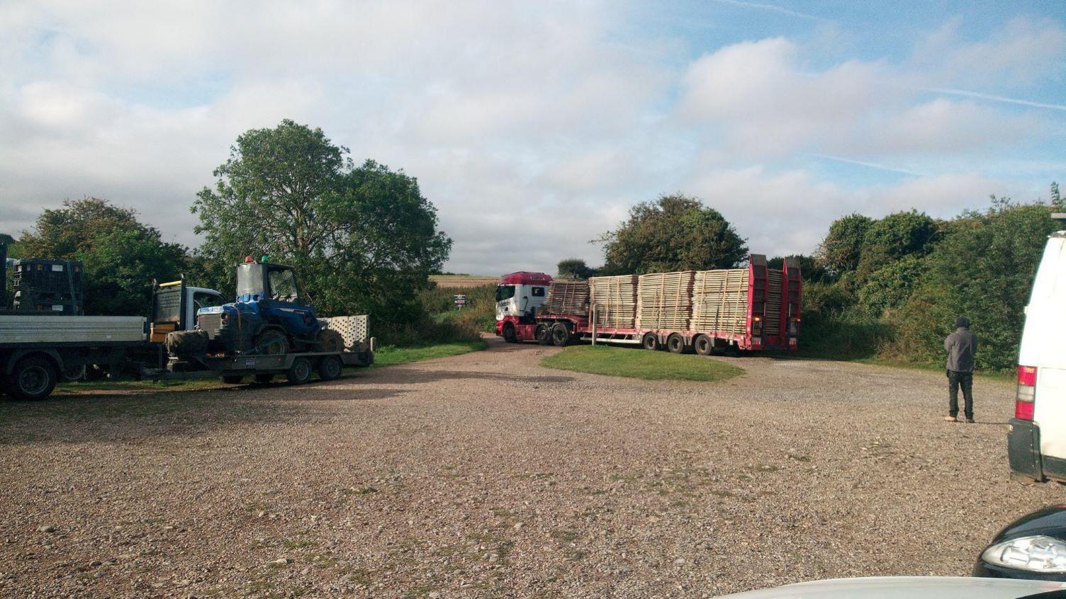 Lorries arriving at Stiffkey Marshes. One lorry is carrying a tractor while the other is carrying wooden boards. They are parked on a gravel path with trees in the background.