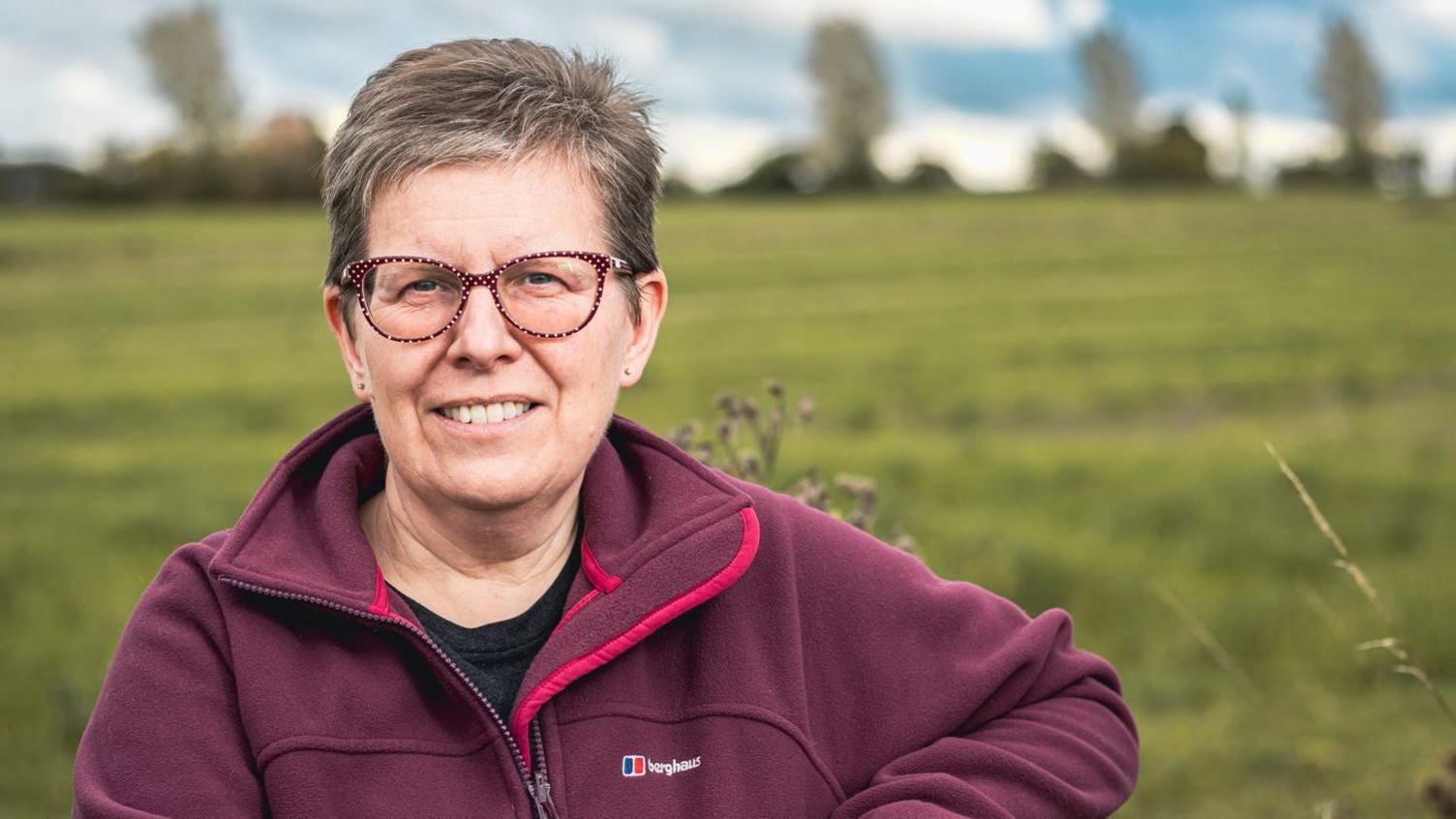 Lorna Vyse smiling at the camera. There is a field in the background. She is wearing glasses and a red fleece jacket with "Berghaus" branding on it