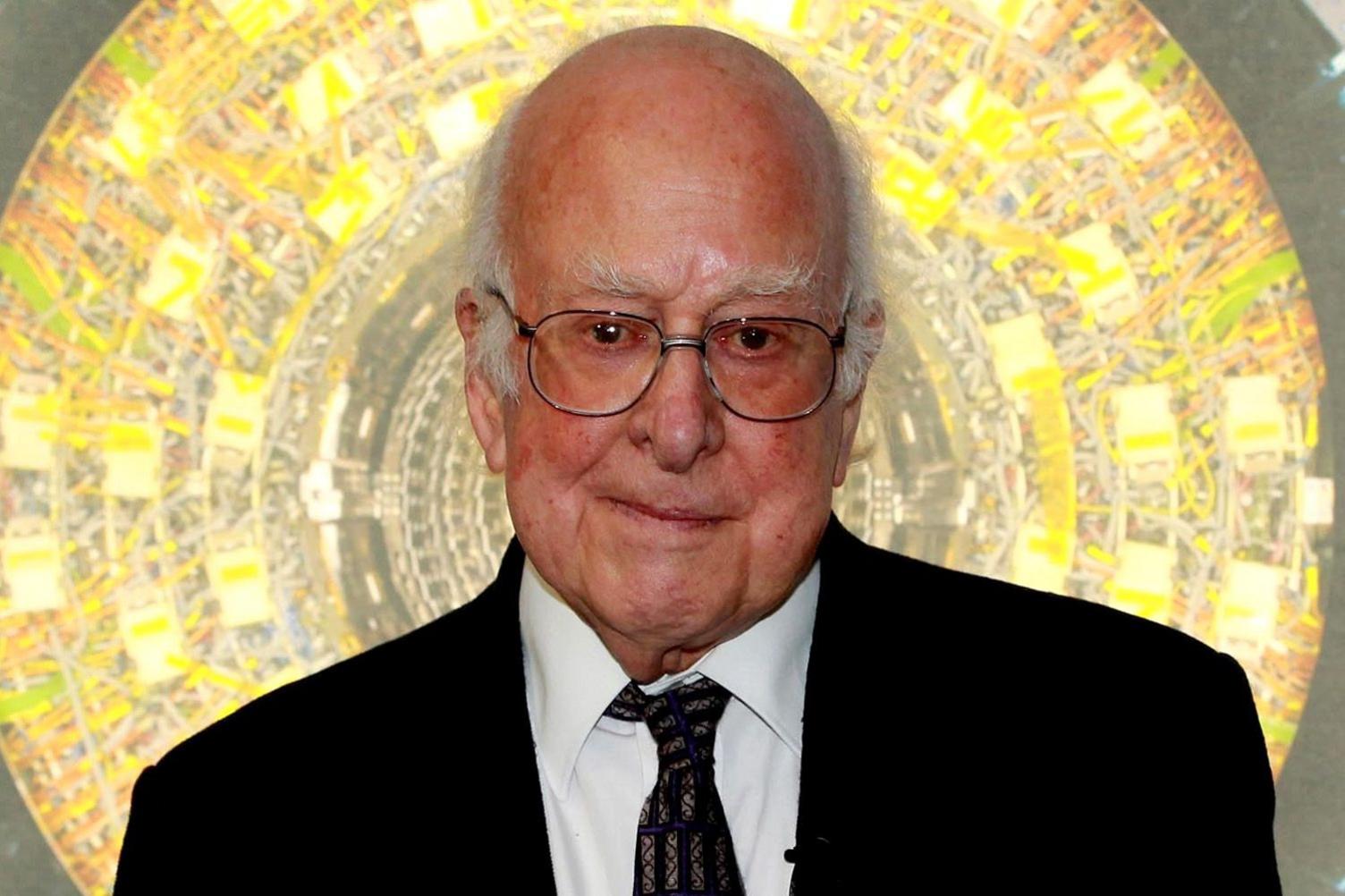 Peter Higgs is photographed in front of what looks like an image of the inside of the Large Hadron Collider. He is an elderly man, bald with bushy eyebrows and glasses. He wears a black suit and white shirt, with. a dark tie slightly askew.