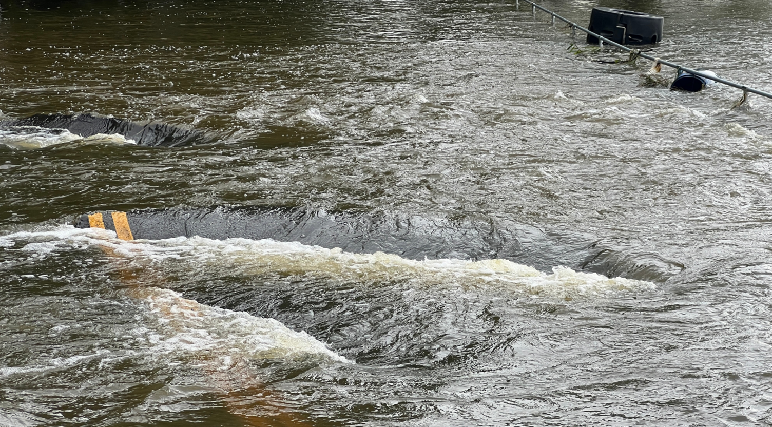 An image of Mill Lane fully submerged under water. There is a chunk of tarmac with double yellow lines on sticking out of the water.