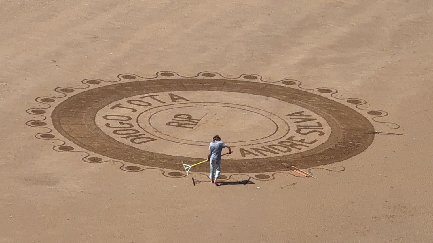 A large and intricate sand art design on a sandy beach. The artwork has multiple concentric circles and detailed patterns. There is a man working on it with a rake.
