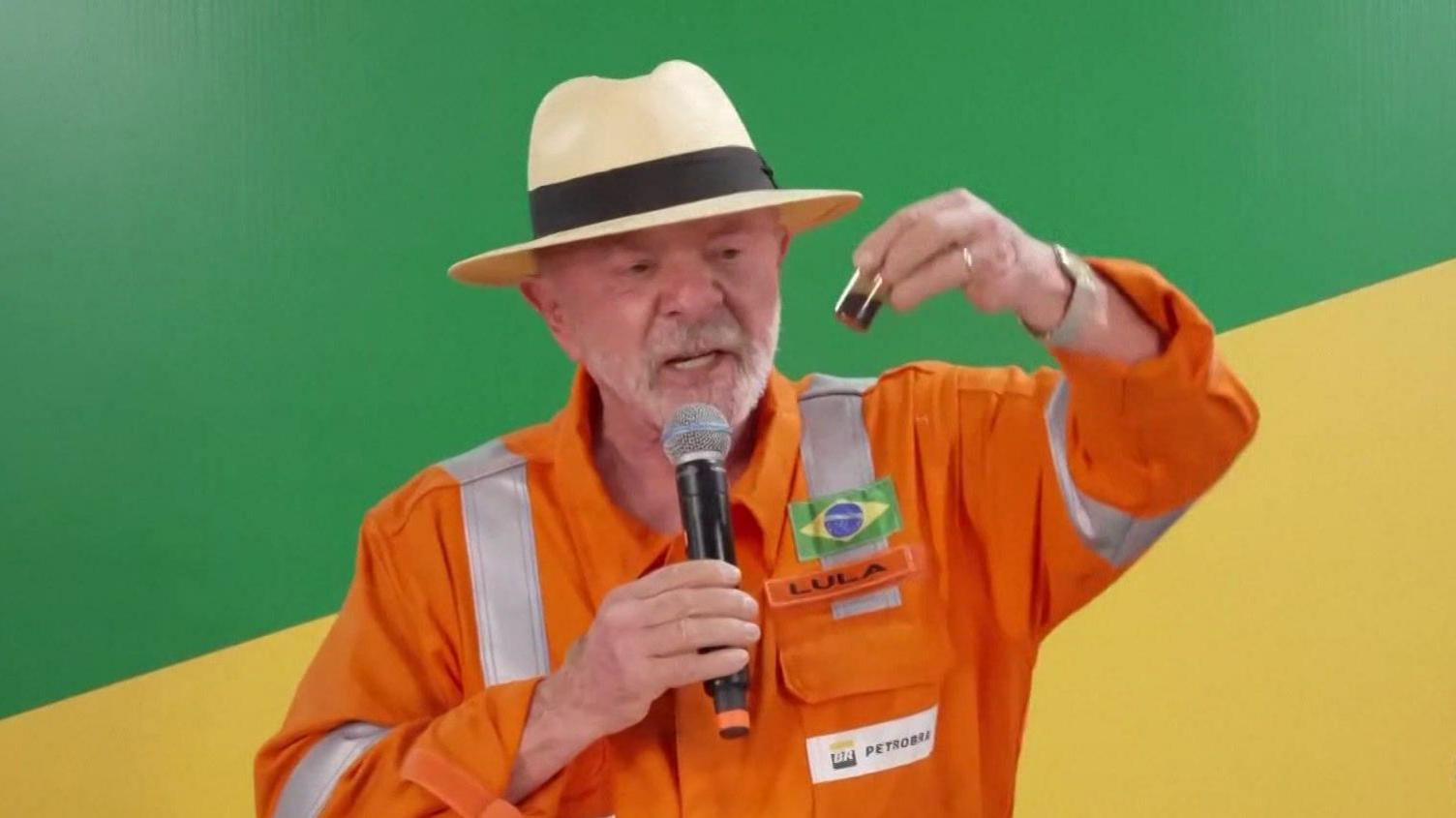 President Lula, wearing an orange jumpsuit with the logo of the state-run oil company Petrobras, hold up a small container filled with oil. He is wearing a cream-coloured Panama hat with a black ribbon around it. In his right hand, he is holding a microphone.