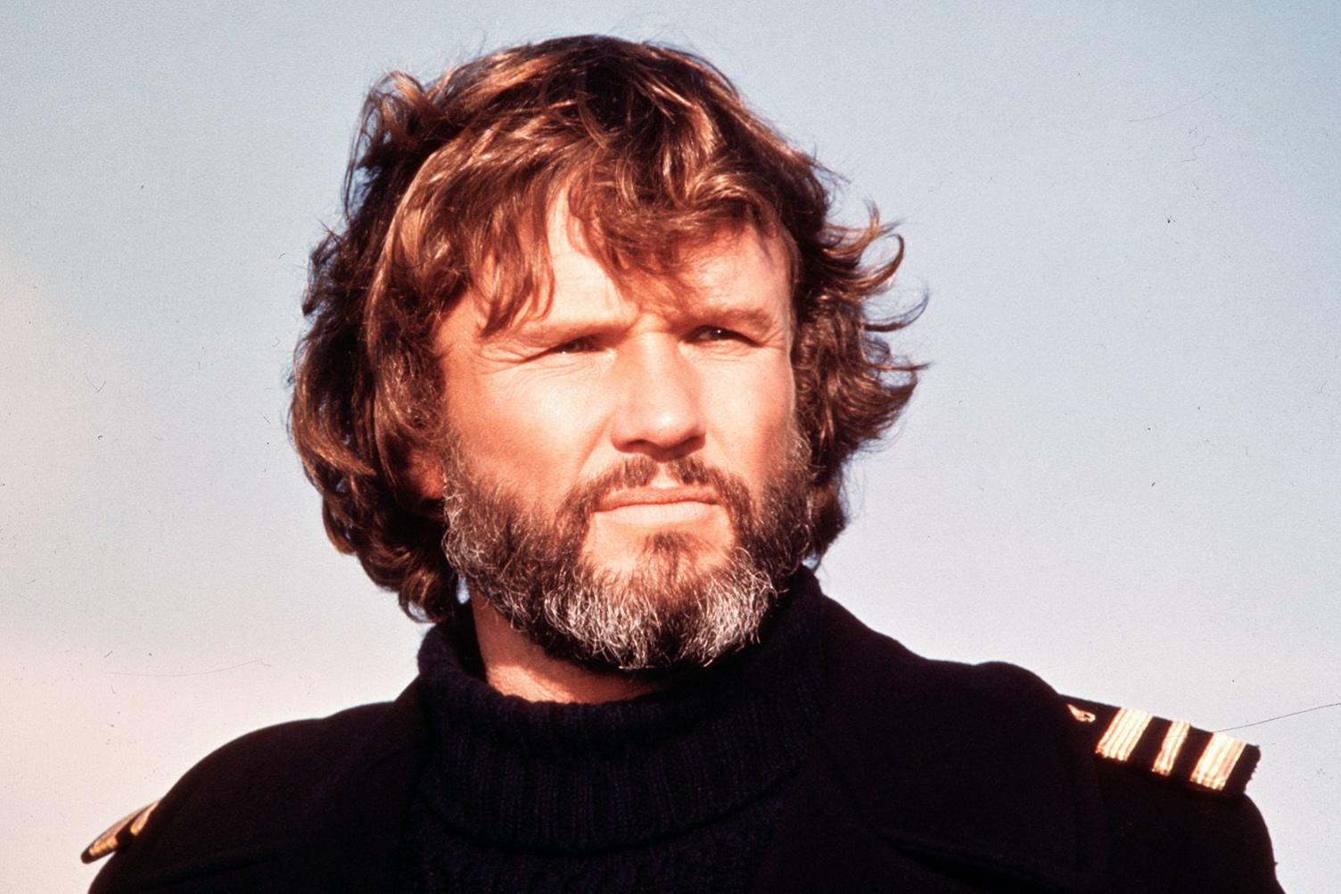 Film still of Kris Kristofferson, taken in the 1970s against a background of sky - he has reddish hair, a greying beard and is wearing a black turtle-neck sweater with epaulettes