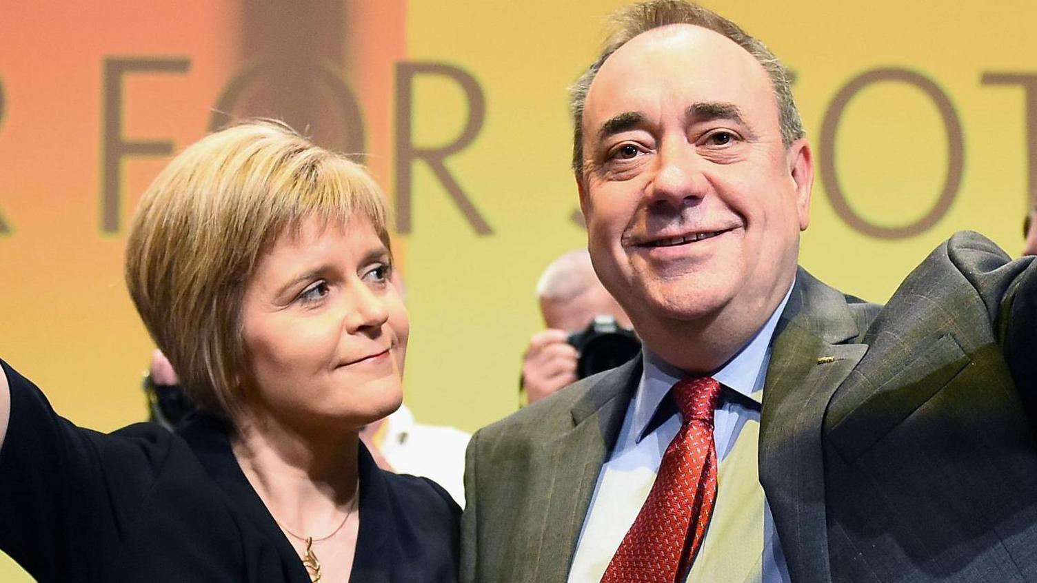 Nicola Sturgeon, who is wearing a black dress, looks at Alex Salmond, who is wearing a grey suit jacket, a blue shirt and a red patterned tie. The two are waving to crowds at the SNP Conference, though the image is cropped in on their faces.