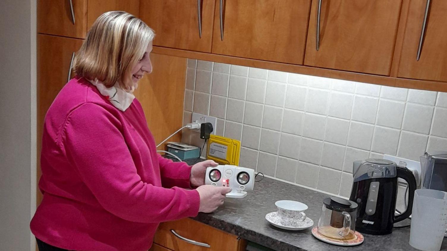 A woman is standing in a kitchen using an mp3 speaker with built-in magnifying lenses. She is smiling, has light shoulder-length hair and is wearing a white scarf and a pink jumper. The kicthen comprises of wooden overhead cabinets, light grey tiles, black speckled work surfaces and a black kettle. A cup and saucer is beside the kettle and a yellow envelope is also sat on the counter.