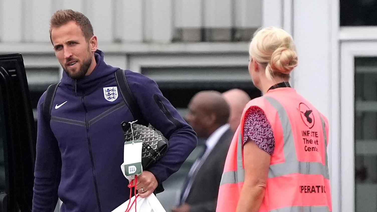 Harry Kane is wearing a navy England tracksuit top and is holding a dark wash bag. He is walking in front of the terminal building at Stansted Airport. To his left is a woman wearing a pink hi-vis jacket.