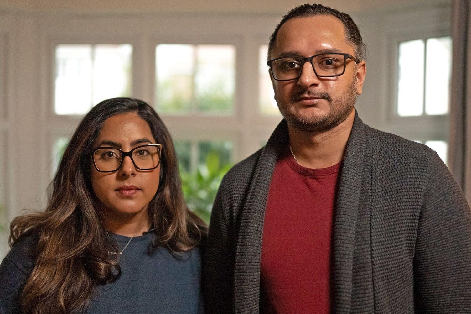 A couple stand next to each other with serious expressions. The light shines through the window behind them.