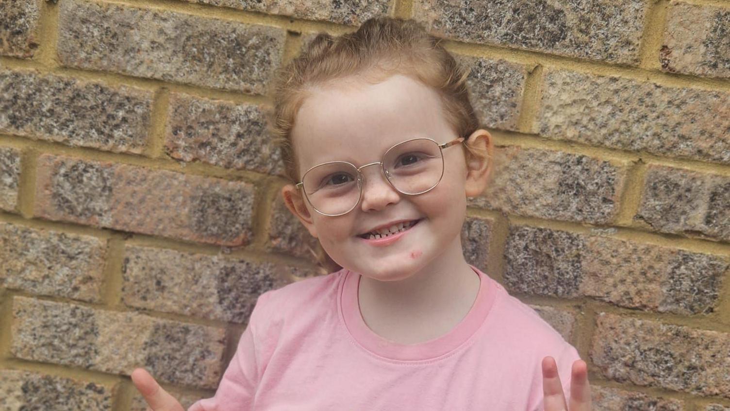 Phoebe is standing in front of a brick wall. She has ginger hair in a ponytail and is wearing a pink T-shirt. She is holding her thumbs up and smiling. 