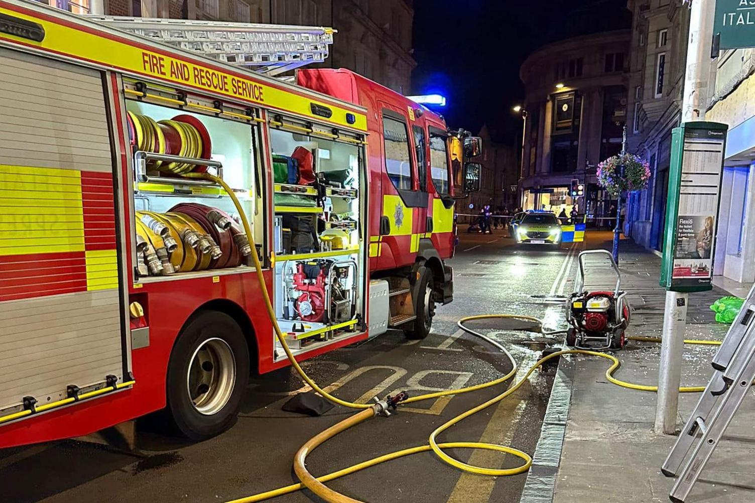 A fire engine on George Street with fire hoses extending onto the street and out of shot. Smashed glass is on the pavement. A police car is in the background.