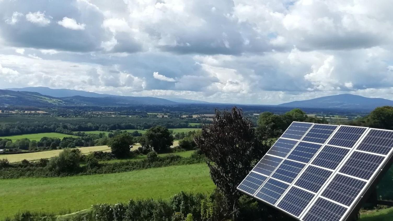 A board of solar panels set on a sloped garden outside the Wilkinson family's home on a sunny day in County Kilkenny. The board can be swivelled to point the panels at the sun's rays.  The garden is surrounded by the green fields of the Suir Valley with hills in the distance.