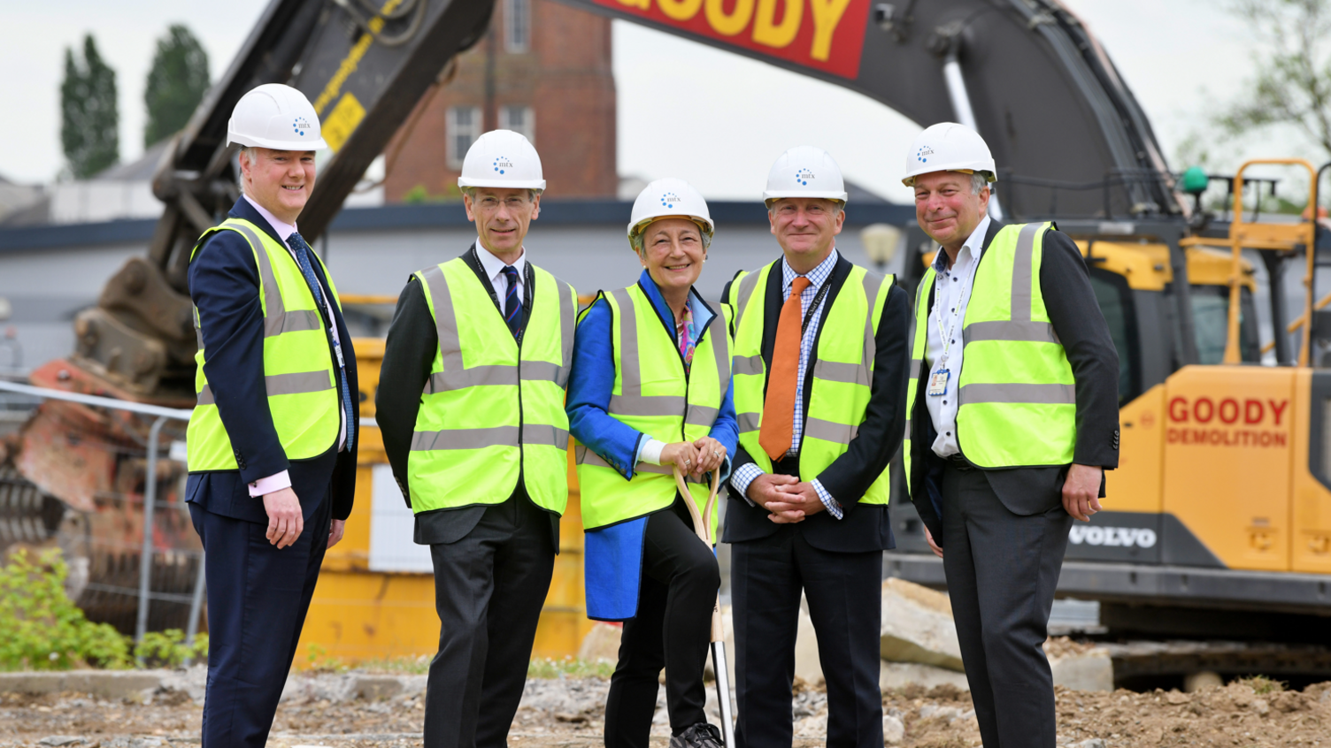 Four men and one woman wearing high-vis and white hard hats stand on a building site. All are smiling. Earth-moving equipment can be seen behind them.