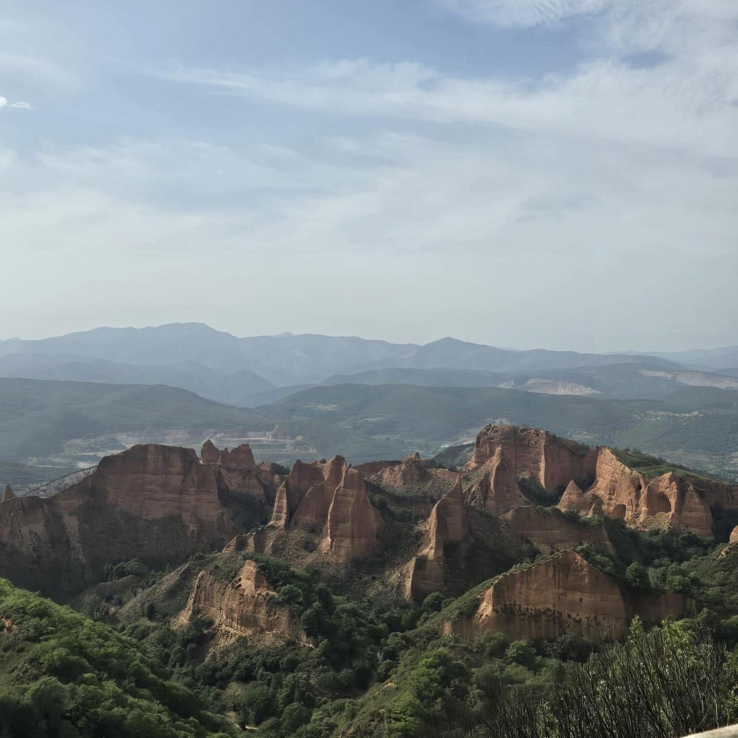 Rock formations with steep, vertical cliffs and pinnacles dominate the foreground of a wide valley landscape. Areas of green vegetation surround the formations, with layered hills and mountains extending into the distance. Thin cloud cover fills much of the sky above the ridge line.